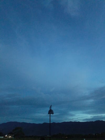 The image depicts a dark blue sky with faint clouds, transitioning into twilight. In the foreground, a silhouette of a mountain range is visible against the horizon. To the right, a solitary, dark, pole-mounted object with a domed top and intricate lattice work is silhouetted against the sky. In the far left middle ground, a building with illuminated windows is partially visible behind dark vegetation. The scene is set in an outdoor environment, likely a field or open area, with a dark, unidentifiable ground cover. The image appears to be taken in Meunasahmanyet, Indonesia, during twilight.