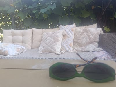 This is a medium-shot, eye-level, outdoor photograph of a patio area with a wicker-style sofa adorned with several textured, neutral-colored cushions, including one with a diamond pattern and tassels, and another with a cross-stitch pattern. The sofa is set against a backdrop of lush green foliage, likely from a vine or climbing plant, with some sunlight filtering through. In the foreground, a pair of green-tinted sunglasses with tortoise-shell arms rests on a light-colored wooden surface, possibly a table. The scene is tranquil and appears to be a comfortable outdoor seating area. The location is identified as Serravalle, Switzerland.