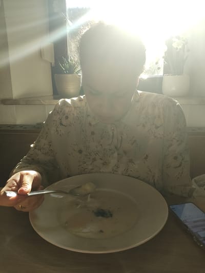 An indoor scene depicts a person enjoying a meal at a table, bathed in bright, natural light. The central subject is a woman, partially obscured by intense sun glare on the left side of the frame. She has dark, curly hair and is wearing a light-colored blouse with a floral pattern.

In front of her is a white, round bowl containing a creamy liquid, likely soup, with some dark specks (possibly pepper or herbs). Her right hand holds a spoon, poised above the bowl, suggesting she is in the process of eating. A distinctive gold ring is visible on one of her fingers.

The table has a light wooden texture. To the upper right, a mobile phone rests on the table. Its screen is illuminated, showing what appears to be a standard smartphone interface, though specific text or icons are unreadable due to blurriness and distance.

The setting appears to be a dining area, possibly a restaurant or cafe, given the table arrangement and lighting. Strong, direct sunlight streams in from a window to the left, causing significant overexposure and a strong lens flare effect across the top left quadrant of the image. This bright light illuminates parts of the woman's hair and the background. Behind her, near the light source, are potted plants and some white decorative objects.

The abundant sunlight indicates a daytime scene, likely sunny weather. Emotions are not discernible due to the facial obscuration. No clearly readable text is present within the image.