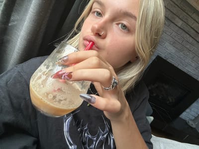 A young blonde female with light eyes and a nose ring in her left nostril is captured in a close-up, chest-up shot, looking directly at the viewer. She is actively drinking from a clear glass filled with a light brown, frothy beverage, using a red plastic straw. Her left hand holds the glass, showcasing long artificial nails adorned with various designs; some are dark, others feature intricate reddish-pink floral patterns. A silver-toned ring with an ornate, possibly gothic, design is visible on a finger of her partially obscured right hand. She is wearing a black t-shirt with a distinctive silver graphic pattern on the chest. The indoor background consists of dark, pleated fabric on the left and a grey brick fireplace structure with a dark, recessed opening on the right. The scene is located in the city of Victoria, Canada.