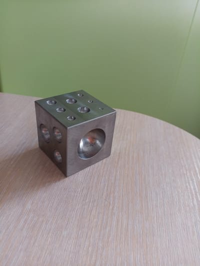 A metal cube with multiple holes is placed on a wooden surface. The cube has a reflective, silver finish and is positioned slightly off-center in the frame. The wooden surface has a light brown, grainy texture with visible wood grain lines running diagonally. The background is a solid, light green color. There are no people or actions depicted in the image. The scene appears to be indoors, likely on a table or desk. The location is not specified as being in Vilyuysk, Russia.