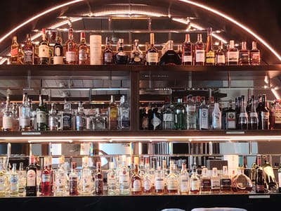 A well-lit, upscale bar features shelves stocked with a wide variety of liquor bottles. The bottles are neatly arranged, with some illuminated by spotlights from above. In the foreground, a dark countertop is visible, reflecting the bottles on the shelf above. The background consists of mirrored panels and lighting fixtures that create a sophisticated ambiance. The scene suggests a high-end establishment, possibly a hotel bar or a dedicated cocktail lounge. There are no people or ongoing activities depicted in the image. The location is identified as Konigil, Uzbekistan.