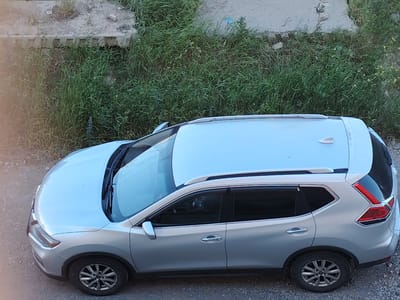 The image, taken from a high angle, shows a silver SUV parked on a gravel surface adjacent to a patch of overgrown grass and a concrete embankment. The car is oriented diagonally, with its front facing towards the bottom left of the frame. The vehicle appears to be a Nissan Rogue, recognizable by its distinctive taillight design and roof rails. The grass is tall and unkempt, suggesting an undeveloped or neglected area. The concrete structure appears to be a retaining wall or part of a larger construction. The overall scene suggests an outdoor location, possibly on the outskirts of a city, consistent with the Border Gyumri-Akhuryan, Armenia area. There are no people visible in the image.
