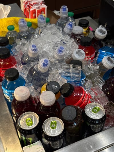 A high-angle shot shows a metal tub filled with ice and various beverages. Bottles of water, Powerade, and "Gold Peak Real Brewed Tea" are mixed in with cans of "Monster Energy" and "Minute Maid" drinks. A yellow towel rests to the side, along with some red and white packaged items. The drinks are surrounded by ice cubes, which suggest a cool or refreshing environment. The location appears to be a beverage cooling station inside an establishment and there is no information on whether this is in Honolulu, United States of America.
