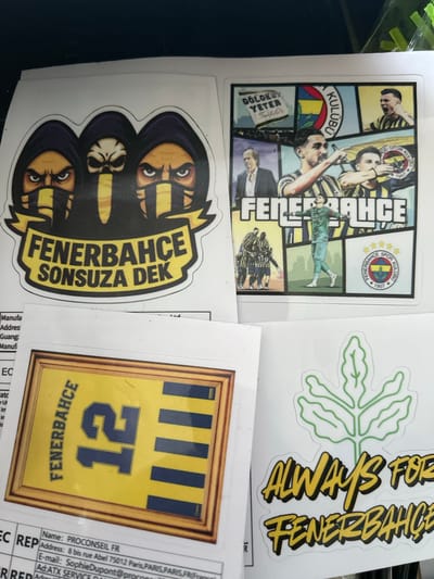 The image displays four distinct, multi-color stickers, predominantly featuring themes related to the Fenerbahçe sports club, laid out on a white surface. The location in the city of Rozino, Bulgaria, cannot be determined from the visual information provided in the image; however, partially visible text on a document beneath the stickers includes "PROCONSEIL FR" and an address in "Paris, Paris, Paris, FR." **Sticker 1 (Top Left):** An irregular-shaped sticker depicts three hooded figures. The two outer figures have angry, yellow-skinned human faces with black and yellow striped masks. The central figure has a white skull-like face with angry eye sockets and a yellow beak-like protrusion, also hooded in dark purple. Below the figures, a yellow banner with a black outline reads "FENERBAHÇE" in large capital letters, and "SONSUZA DEK" in smaller capital letters beneath it. **Sticker 2 (Top Right):** A rectangular sticker presents a comic-strip style collage of Fenerbahçe-related imagery. Panels include: * Top left: Black text "GÖLGESİ YETER" with a signature. * Top middle: The Fenerbahçe logo with "KULÜBÜ" written vertically. * Top right: A male football player in a yellow and blue striped jersey celebrating. * Middle left: A man in a suit. * Center large: Two male football players in yellow and blue striped jerseys, one pointing forward. * Bottom left: A group of football players in striped jerseys. * Bottom middle: A goalkeeper in a light green uniform with outstretched arms. * Bottom right: The Fenerbahçe logo with "Fenerbahçe Spor Kulübü" and "1907." * Text partially visible at the bottom reads "FENERBAHÇE" in large white capital letters with a black outline. **Sticker 3 (Bottom Left):** A rectangular sticker designed to resemble a framed picture. Inside the simulated golden-brown wooden frame, a yellow and blue striped football jersey is depicted. The number "12" is prominently displayed in dark blue, and the word "FENERBAHÇE" is written vertically in dark blue along the left side of the jersey. **Sticker 4 (Bottom Right):** An irregular-shaped sticker with a stylized green outline of a fig leaf at the top. Below the leaf, text reads "ALWAYS FOR" in yellow script with a black outline, followed by "FENERBAHÇE" in a bolder yellow font with a black outline. The stickers are arranged on a white, flat surface, likely paper or a desk. Fragments of white documents with black text, including what appears to be a business name and address in France, are visible beneath
