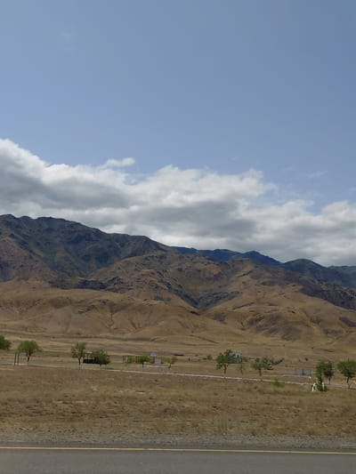 The image depicts a dry, mountainous landscape with sparse vegetation. In the foreground, a paved road with a yellow dividing line runs horizontally across the frame. Beyond the road, a field of dry grass stretches towards a row of scattered trees and some small, man-made structures, possibly picnic areas or small buildings, spaced along a white line. In the mid-ground, rolling hills are covered in dry, golden-brown grass. The background is dominated by a range of rugged mountains, with varying shades of green and brown, indicating some vegetation on their slopes. The sky above is a light blue with scattered, puffy white clouds and some darker, grey clouds. The overall scene suggests a rural, possibly arid environment.