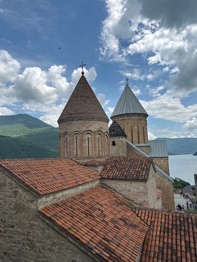 The image shows a view of the Ananuri fortress complex in Shalikiantkari, Georgia. The foreground features a close-up of several terracotta tiled roofs belonging to the fortress buildings. In the middle ground, two prominent church towers rise above the roofs. The left tower is conical and made of brick with arched windows, topped with a cross. The right tower is taller and has a metal conical roof, also topped with a cross. Between these two towers, a smaller, darker structure with a pointed roof is visible.
In the background, a body of water, likely the Zhinvali Reservoir, stretches out. Beyond the reservoir, lush green, forested mountains rise against a partly cloudy blue sky. Several people can be seen walking along a path on the right side of the image, near the water's edge, and a few small birds are flying in the distance. The architecture suggests a historical religious site.