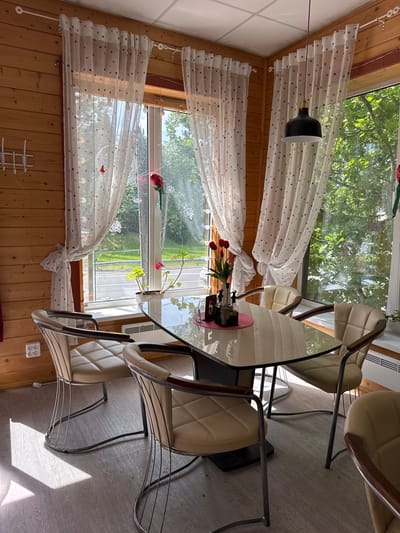 The image shows a dining area with a glass-top table and four chairs. The room has wooden walls and a large window with sheer, polka-dot curtains. A black pendant light hangs from the ceiling. On the table, there is a vase with red tulips, a coaster, and some condiment bottles. Outside the window, trees and a road are visible. The scene appears to be a cafe or restaurant in Serovo, Russia.