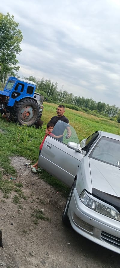 In Chernyavskii, Russia, on a grassy field with a dirt path and a backdrop of deciduous trees under a cloudy sky, a man and a child are by a silver sedan with its driver's side door open. The man, wearing a black t-shirt, stands with his right arm around the child. The child, wearing a red shirt and green clogs, is being held by the man. To the left of the scene is a blue tractor, with the inscription "T-34" visible on its side. The car's hood is partially open, and the driver's side door is ajar. The ground is a mix of dirt and grass.