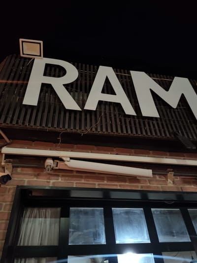 A nighttime exterior shot captures a large white sign spelling "RAM" against a dark sky and a corrugated metal facade. Below the sign, a brick wall supports a row of windows and a security camera. A floodlight is mounted on the roof above the sign, illuminating the letters. The lower portion of the image shows reflections in the glass windows. The scene is likely an establishment in the city of Ariana, Tunisia, given its architectural style and the presence of a sign that could indicate a business.