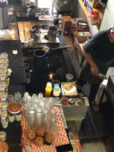 A high-angle, full shot captures a busy kitchen environment with a person in the foreground. The subject, wearing a dark shirt and apron, is partially visible, with their hands engaged in an unseen task. The scene is cluttered with various kitchen items, including numerous cans of food arranged neatly on shelves and a counter. Several stacks of clear drinking glasses and brown ceramic cups are also prominent. Metal pots and pans, some steaming, are present on a stove. In the background, shelves hold boxes of what appear to be food products and other supplies. The overall impression is one of a working kitchen, possibly a restaurant or food preparation area, characterized by its functional and somewhat industrial aesthetic.