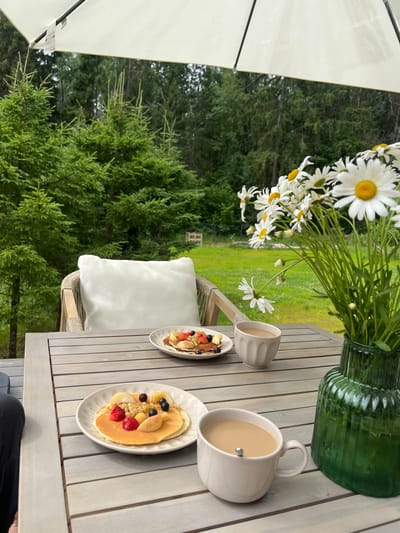 The image depicts an outdoor table setting with two plates of pancakes topped with various fruits, two cups of coffee, and a vase of daisies. The table is made of light-colored wooden planks and is situated on a deck, likely in a residential setting. In the background, there are evergreen trees and a grassy area, suggesting a natural or park-like environment. The overall scene conveys a sense of a relaxed breakfast or coffee break in a pleasant outdoor setting. The location is identified as Popovka, Russia.