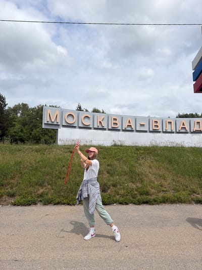 A young woman wearing a pink baseball cap, a white t-shirt, and a plaid shirt tied around her waist, along with light green sweatpants and white sneakers, stands on an asphalt surface. She is holding a pink smartphone in her right hand, seemingly taking a selfie, and a red rope, possibly a jump rope, dangles from her left hand. The woman is positioned in front of a large sign with large, light orange letters spelling out "МОСКВА-ВПАД" against a grey background, situated on a grassy embankment. Behind the embankment, a line of trees forms the horizon under a cloudy sky. The sign appears to be a directional marker, with a Russian flag motif visible to the far right. The environment suggests an outdoor location on the outskirts of Nikolaevka, Russia, possibly marking an entrance or administrative boundary.