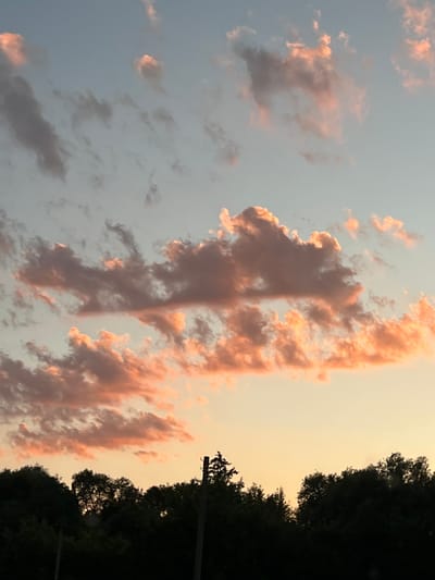 The image depicts a sky at dusk, with clouds illuminated by the setting sun. The sky transitions from a pale blue at the top to a warm orange hue near the horizon. Scattered clouds, some tinged with pink and orange, are visible against the sky. Silhouetted against the sky is a dense line of dark green trees, forming the treeline of a forest. A tall, thin, vertical pole, possibly a utility pole, stands prominently on the right side of the frame, with two horizontal fixtures near its top. The overall scene appears to be an outdoor landscape, potentially on the outskirts of a populated area, given the presence of the pole. The location is identified as Tovstoluh, Ukraine.
