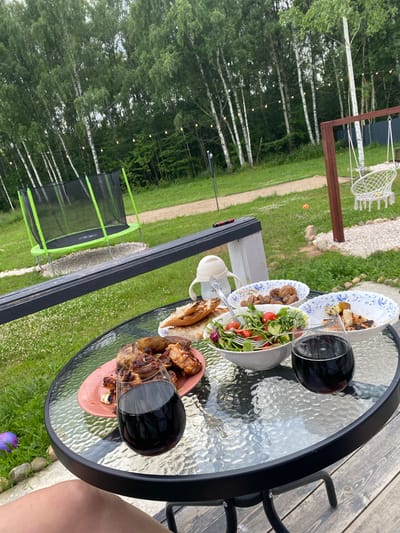 The image shows a glass table laden with food and drinks outdoors in what appears to be a backyard in Ryzhkovo, Russia. A person's leg is visible in the foreground. On the table are two glasses of red wine, a plate of grilled meat and potatoes, a bowl of salad with tomatoes and cucumbers, and another bowl of cooked mushrooms. In the background, a trampoline and a swing set are visible, suggesting a recreational area. The environment is lush and green, with birch trees lining the background. String lights are hung between the trees, indicating it might be evening.