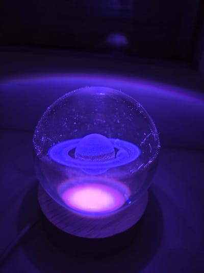 A glass sphere containing a blue, ringed object is illuminated with purple and pink light. The sphere sits atop a wooden base. The background is dark and indistinct.