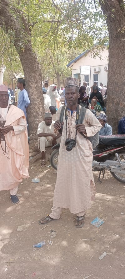 An outdoor scene in Sani Mai Nagge, Nigeria, depicts a public gathering area under bright daylight, with dappled shade from numerous trees with green foliage and visible trunks.
In the foreground, centered, a male adult, appearing middle-aged with a beard, stands facing the camera. He wears a light-colored, patterned traditional tunic (caftan) and matching trousers, a patterned cap, and dark-colored flip-flops. A black backpack is on his shoulders, and a black Nikon DSLR camera with a lens is suspended around his neck by a black strap bearing the "Nikon" logo. His hands are positioned near the backpack straps.
To the immediate left of the central subject, a partially cropped male adult, also in traditional attire including a light peach-colored tunic and cap, wears dark blue shoes and holds dark, beaded objects. Behind them, numerous other individuals, both male and female adults, are dispersed throughout the background. Men are generally seen in various colored traditional tunics and caps, while women are observed wearing head coverings, some in hijabs. One male adult is seated on the dirt ground beneath a tree, wearing sandals.
The ground consists of dry, uneven dirt, with scattered refuse including plastic bags, paper, and other small debris. A black and silver motorcycle, resting on its kickstand, is partially visible on the right side behind the central subject. In the far background, a light-colored building with a blue-sloped roof, exterior air conditioning units, and an unreadable wall sign is present.
