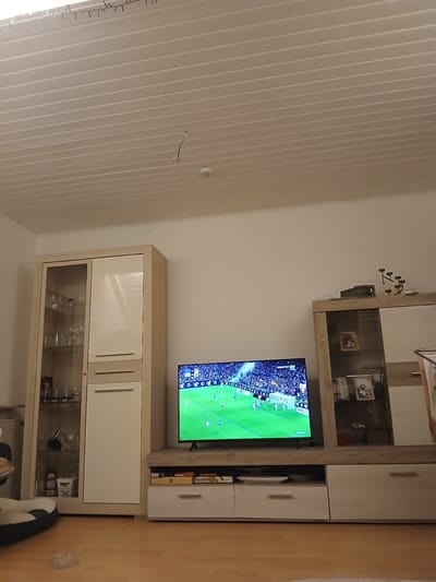 The image shows a living room interior in a home that is located within the city of Homburg, Germany. A large, built-in entertainment center dominates the view, featuring a large-screen television displaying a football match. The entertainment center includes glass-fronted cabinets, wooden-framed cabinets, and drawers. Several objects are visible within the cabinets, including glassware. The room features a white, paneled ceiling. A partial view of the flooring is seen on the right. There are items positioned in front of the unit, including shoes. The surrounding environment consists of the walls and floor of the room.