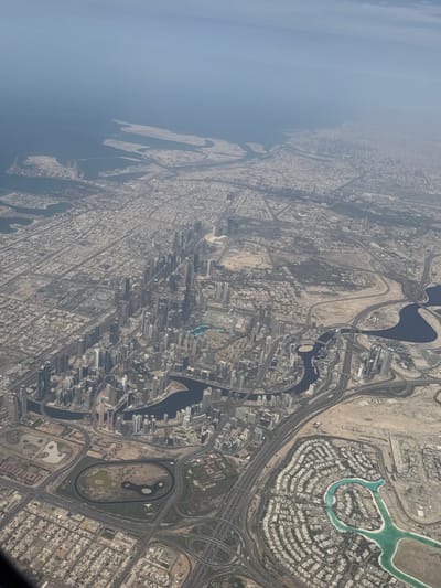 This is an aerial image of Dubai, United Arab Emirates, captured under a hazy sky. The upper-left quadrant distinctly shows the Deira district, characterized by a dense, gridded urban layout extending to the coastline. Offshore from Deira, within the Arabian Gulf, several large artificial landforms associated with the Deira Islands project are visible.
The central and lower portions of the image display a vast, modern urban expanse. A prominent, meandering waterway, identified as the Dubai Water Canal/Creek, carves through the cityscape, flanked by a high concentration of contemporary skyscrapers and multi-story buildings that form a central business and residential corridor. An intricate network of multi-lane highways and extensive, multi-level interchanges connects various parts of the city.
In the lower-right quadrant, curvilinear residential developments featuring distinctive artificial turquoise lagoons are observed. Towards the lower-central area, a large, dark oval-shaped track and other green, manicured spaces, likely part of a sports complex or golf course, are discernible. Patches of undeveloped sandy terrain are interspersed among the urban developments. No individual people or specific actions are resolvable due to the high altitude perspective.