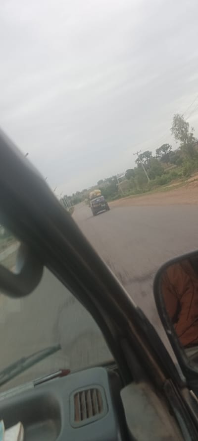 The image shows a road in the city of Barakin Kuru III, Nigeria. The photo was taken from inside a moving vehicle. Another car, with items piled on its roof, is visible further down the road. The sky is overcast. The side view mirror is visible. Trees line the roadside.