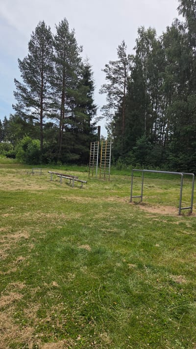 This is a medium shot, eye-level, outdoor photograph taken on a cloudy day at a park in Saurieši, Latvia. The foreground features a grassy field with dry patches of straw and sparse white clover flowers. In the middle ground, several pieces of outdoor exercise equipment are visible. To the left, there are low parallel bars and a bench with a wooden seat and metal supports. Further to the right are two tall, ladder-like structures made of metal, one with yellow accents. On the far right, a set of parallel bars for exercising stands on a patch of sand. The background is dominated by a dense line of tall, mature pine and birch trees. The sky is overcast. No people are present in the image.