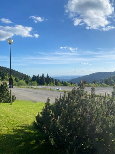 The image shows a partially cloudy sky with some land. On the right is a large evergreen tree. A paved road runs through the landscape, with a white car parked on the side. In the distance, a mountain range is visible. A street lamp is in the foreground on the left. The location is likely near Loučná nad Desnou, Czechia, given the landscape and the presence of mountains.