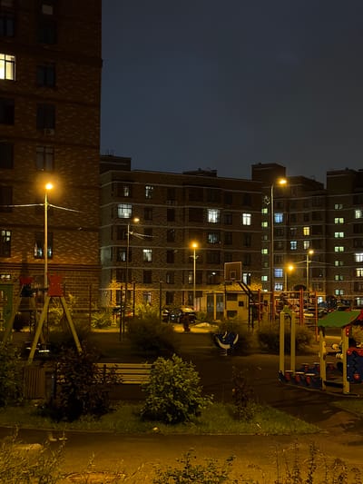 The image depicts a nighttime view of a residential courtyard in Saburovo, Russia, specifically showing an apartment complex and playground. The sky is dark blue, suggesting evening or early night. Several multi-story apartment buildings, constructed with brickwork, dominate the background. Lights are visible in many of the windows, indicating occupancy. Streetlights illuminate the courtyard, casting a warm glow on the scene.

In the foreground, a children's playground is visible. It includes a swing set with red supports, a basketball hoop with a backboard, and various other playground equipment with colorful components. There is also a park bench and a trash receptacle. Greenery, including bushes and grass, is present around the playground and the buildings. The ground surface appears to be asphalt or a similar paved material, with some areas wet, possibly from rain or dew. The overall impression is of a well-lit, residential area at night. There are no people visible in the image.