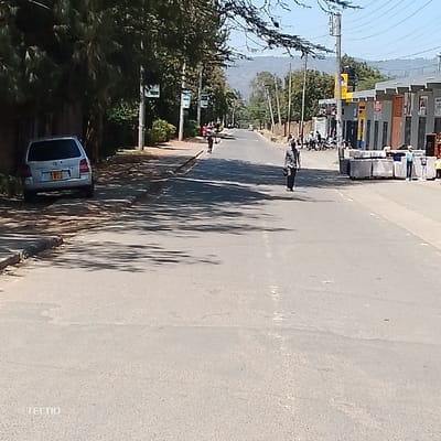 The scene depicts a daylight street view in Machakos Central ward, Kenya. A two-lane asphalt road, lacking visible lane markings, extends from the foreground into the distance. Strong sunlight casts distinct shadows of overhead trees and objects onto the road surface.
On the left side of the road, a silver five-door SUV with Kenyan license plate KBR 191S is parked on a paved shoulder, facing away from the viewer. Behind the vehicle is a dense line of green trees and foliage, partially obscuring a perimeter wall. Further along this side, a paved sidewalk is visible, bordered by additional trees and utility poles carrying advertising banners. Several individuals are observed walking along this sidewalk in the mid-distance.
On the right side of the road, a commercial area features several single to two-story buildings with dark-framed windows and doors, some equipped with awnings. In the foreground, numerous rectangular objects, possibly building materials or mattresses, are stacked in rows on the paved shoulder adjacent to these establishments. Several individuals, including two small children, are present near these stacked objects. Further down the street, a group of people and multiple motorcycles or scooters are gathered in front of the commercial buildings.
In the center of the roadway, a person wearing a dark top and light pants walks away from the camera. Another individual is visible walking further down the road.
The background features rolling hills or mountains covered in vegetation under a clear, light blue sky. Utility poles line both sides of the street extending into the horizon. A "TECNO" watermark is present in the lower-left corner of the image.