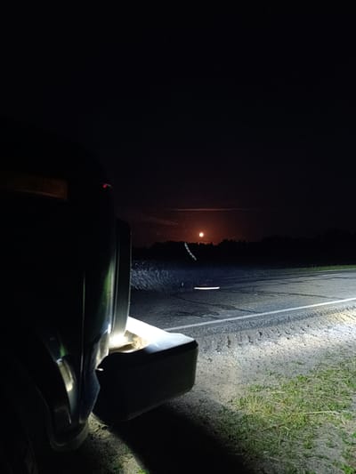 This image depicts a nighttime scene on a rural road, possibly in the vicinity of Мёрскі сельскі Савет, Belarus. The perspective is low, looking from the rear of a dark-colored vehicle, with its headlights illuminating the asphalt road and the grassy shoulder. In the distance, silhouetted against a dark sky, is a forest line. A bright, orange-hued moon is visible in the sky, casting a soft glow. The road appears to have some wet patches or debris, and there are faint light streaks that could indicate movement of other vehicles or light sources. The overall atmosphere is dark and somewhat isolated.