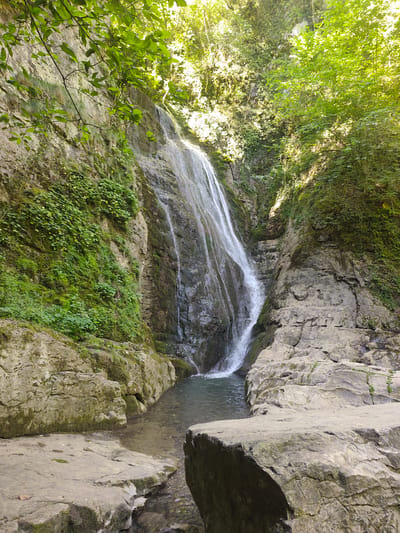 A scenic view of a waterfall cascading down a rocky cliff face, surrounded by lush green vegetation and trees. The water forms a pool at the base before flowing into a shallow stream. Large, weathered rocks are visible in the foreground and along the stream bed. The scene is bathed in natural sunlight, highlighting the textures of the rocks and the vibrant greenery. The location is identified as a waterfall near the town of Teteven, Bulgaria.