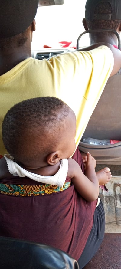 An overhead view within a vehicle in Ijotan, Nigeria, shows an adult and a child from behind. In the midground, an adult, likely of African descent, wears a yellow short-sleeved top and is seated in what appears to be a front passenger or driver's seat, facing forward. On the adult's front, a baby or young child, also of African descent, is positioned, facing forward. The child has closely cropped dark hair and wears a white, scalloped-edge undershirt. A maroon fabric with a teal, yellow, and black geometric pattern wraps around the child's back. The child's right arm and bare feet are visible, with hands clasped in front. To the immediate foreground, the back of another individual's head, wearing a black cap, is visible, suggesting a third person is seated behind the adult and child. In the background, visible through the vehicle's interior, are dashboard vents, a portion of a seat frame, and a clear plastic bottle with a red label (possibly a soft drink) resting in a door compartment. Bright daylight is visible through the windshield.
