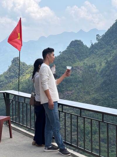 Two people stand on a platform overlooking a mountainous landscape in Lũng Phìn Commune, Vietnam. The man, wearing a long-sleeved white shirt and jeans, holds up a plastic cup with a dark liquid. The other person, partially obscured behind him, has long dark hair. A Vietnamese flag is positioned to the left of the scene. A metal railing borders the platform, and chairs are partially visible at the edge. The background features lush green mountains and a partly cloudy sky.
