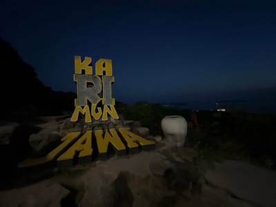 A large, three-dimensional sign in the foreground reads "KARIMUNJAWA." The letters are stylized and appear to be made of concrete with yellow detailing. The sign is set against a dark blue night sky, suggesting the image was taken after sunset. To the left of the sign, a dark, silhouetted hill or mountain slopes downward. To the right of the sign, a large, white ceramic pot sits on the ground. The surrounding area is dimly lit, with some vegetation and rocky terrain visible. In the distance, faint lights suggest a settlement or town, possibly on an island, with a body of water between the foreground and the distant lights. The overall mood is serene and peaceful, with a hint of mystery due to the low light conditions. The location is identified as Karimunjawa, Indonesia.