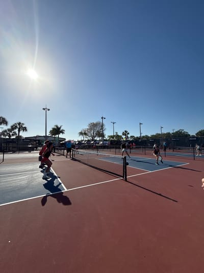 This media file captures an outdoor pickleball match in progress. The scene is set on a two-toned court, featuring blue and reddish-brown sections, with a white net dividing the playing area. Several individuals are visible; two players are prominent near the net on one side, one of whom is captured mid-jump with a paddle extended, seemingly hitting a ball. Another player is positioned further back on the same side of the court. Long, distinct shadows of players and light poles stretch across the court, indicative of either early morning or late afternoon sun. The sun's intense glare is visible in the upper left corner of the frame, confirming a bright, clear day.
In the background, beyond the court, there are numerous tall light poles, black chain-link fences, and a line of trees, including several palm trees, suggesting a warm climate. More people are scattered in the background, possibly spectators or other players. Visible on the black fence is the text "Franklin X-40," which is a brand of pickleball. The setting is an outdoor sports facility, located in Naples, United States.