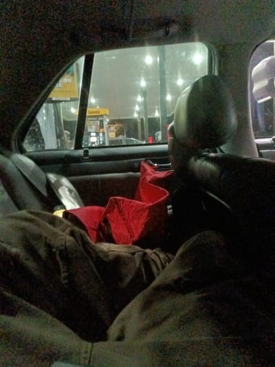 The interior of a car is shown, with a passenger presumably reclining in the back seat. A red garment is crumpled on the seat. Outside the window is a brightly lit gas station with the "Love's" logo visible. The scene appears to be at night or during a time of low light. The interior of the car is dark, and only the illuminated gas station provides external light.