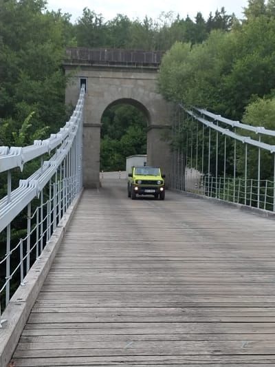 A bright yellow, four-door Suzuki Jimny is driving across a wooden suspension bridge. The bridge has white metal railings with thick cables running along the sides. The bridge spans an archway made of stone. Trees and foliage line the sides of the bridge, and a dense forest is visible in the background. The sky is overcast. The license plate on the car reads BC7 5132. The scene appears to be located in the countryside, with no immediate signs of urban development.