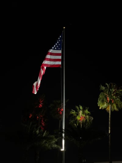 The image displays a United States flag positioned centrally on a vertical flagpole at night in San Bernardino, California. The flag is partially unfurled, hanging downwards, and exhibits its red and white stripes and a blue canton with white stars. It is illuminated from below by an upward-facing light source mounted near the base of the metallic flagpole. Several palm trees are visible in the background against the dark sky. A prominent tall palm tree on the right features green-illuminated fronds with distinct red lighting at the base of its fronds and trunk. Other, less defined palm trees are present, some showing faint red and green illumination. No individuals, vehicles, or dynamic actions are depicted. The scene is static, illuminated solely by artificial light sources.