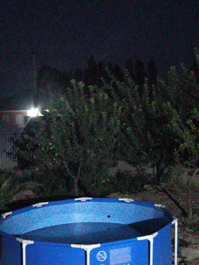 The image captures a scene at night in Cholpon-Ata, Kyrgyzstan, featuring a blue, above-ground swimming pool filled with water. The pool has white support poles and a patterned blue interior. Behind the pool, lush green trees are visible, some bearing small, light-colored fruits. In the background, a bright light source illuminates part of a building and casts a strong beam. The overall environment appears to be a residential or garden area at night.