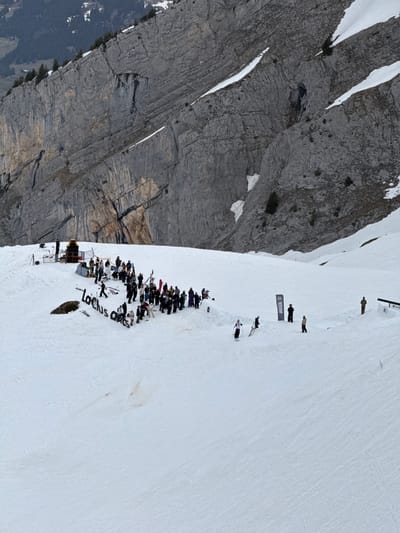 The image shows a gathering of people on a snow-covered slope in La Giettaz, France, located near a rocky mountain face. A crowd is gathered around a small stage or setup with speakers and equipment, with "Lactus Arts" written in the snow. Further up the slope are individuals standing and walking, with a "Return" banner visible. The environment is dominated by the snow-covered mountain slope and the adjacent rocky terrain.