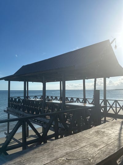 The image depicts a wooden gazebo situated on a pier over a sandy beach and the ocean. The gazebo has a dark, sloped roof and is supported by wooden posts. Inside the gazebo, tables and chairs are visible. The pier itself is constructed from dark wood with a decorative lattice railing. The ocean stretches to the horizon under a clear blue sky with a few wispy clouds. Sunlight is casting shadows on the wooden surfaces, indicating it is daytime. The location is the city of A R A, Indonesia.