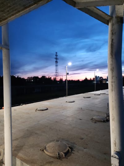 An outdoor, eye-level, medium shot captures a twilight scene over a metallic platform in Sheksna, Russia. The sky transitions from deep blue at the top to hues of pink and orange near the horizon. A tall, latticed transmission tower stands prominently against the colorful sky in the distance, flanked by silhouetted trees and industrial buildings. Two streetlights are illuminated, casting a soft glow on the scene. In the foreground, the concrete platform is marked with several circular manhole covers. The scene is framed by white metal pillars and a section of a metallic roof structure on the left. The overall atmosphere is calm and serene, with the natural light of dusk creating a dramatic backdrop. There are no people visible in the image.