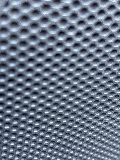 This media file is a close-up, abstract shot of a textured surface. The surface is a uniform grey and features a repeating pattern of small, raised bumps or indentations arranged in a hexagonal grid. The lighting creates strong shadows and highlights, emphasizing the three-dimensional nature of the texture. The perspective is at an angle, with the pattern appearing sharper and more defined in the lower right portion of the image and becoming progressively more blurred towards the upper left. There is no discernible text, people, animals, or specific objects visible. The scene is abstract and focuses solely on the visual characteristics of the textured material. The time of day, weather, and specific location within Parekklisia are not evident from the image itself.