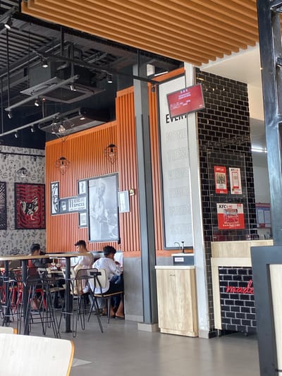 The image depicts the interior of a KFC restaurant, likely in Lhoksukon, Indonesia. The scene is set during the daytime, as evidenced by the bright lighting, though no specific weather cues are visible. The restaurant features a modern design with a mix of wooden slatted walls, black subway tiles, and illuminated signage.
Several individuals are present, primarily seated at tables and engaged in activities that suggest a casual dining or study environment. One group of young men appears to be studying or working on laptops at a table, while others are seated nearby, some looking towards the camera and others focused on their tasks. The overall mood seems relaxed and typical of a fast-food establishment.
Notable details include prominent KFC branding, with logos and promotional posters visible on the walls. A large sign displays the days of the week from Monday to Saturday, likely indicating operating hours or special offers. A red digital display screen is also visible, showing text in what appears to be Indonesian. The restaurant's interior is equipped with various light fixtures, including industrial-style pendant lights. The presence of the menu and seating arrangements confirms it's a food service establishment.
