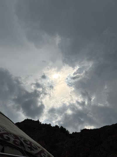 The image depicts a dramatic, cloudy sky with the sun partially breaking through, casting a bright, diffused light. Below the sky is a dark, mountainous landscape, silhouetted against the lighter sky. In the foreground, a portion of a yurt is visible, showing its textured exterior with decorative patterns. The yurt appears to be situated in a rural or mountainous area, consistent with the location of Kyzyl-Korgon, Kyrgyzstan, known for its natural beauty and traditional yurts. The overall impression is one of rugged, natural scenery under a dynamic sky.
