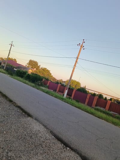 This media file captures an outdoor scene on a clear day, likely in the late afternoon or early evening due to the warm, soft lighting. The setting appears to be a rural or semi-rural area, possibly a residential street in Staronizhestebliyevskaya, Russia.
The primary subjects are utility poles with electrical wires stretching across the frame, a paved road that occupies the lower portion of the image, and a long, reddish-brown fence with brick pillars that runs along the right side of the road. Beyond the fence, glimpses of houses with red roofs and lush green trees suggest a residential neighborhood. The ground on the left side of the road is covered with gravel and small stones.
There are no people or animals visible in the image. No specific activity or event is depicted; it is a static, observational shot of the environment.
Notable details include the gentle slant of the road and the power poles, suggesting the photo was taken from a slightly tilted perspective or a sloped area. The clear sky and the quality of light indicate pleasant weather conditions. The fence and the style of the houses provide location cues consistent with a Russian setting. There is no visible text in the image.