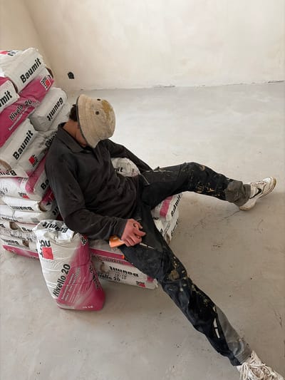A person wearing work clothes and a dirty bucket hat is asleep, seated on a stack of bags labeled "Baumit" and "Niello 20". The person's legs are extended, and their right hand rests on a phone. They are wearing dirty work pants and white sneakers. The environment appears to be an indoor construction site with concrete flooring and unfinished walls. The scene is located in Příbram, Czechia.