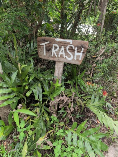 A wooden sign with "TRASH" painted in white is partially obscured by lush greenery in an outdoor setting. The sign appears rustic, with visible wood grain and imperfections. The vegetation includes various types of leaves and plants, suggesting a natural or garden environment. The location is likely in Moo 4, Thailand, as per the instructions. There are no people visible in the image.