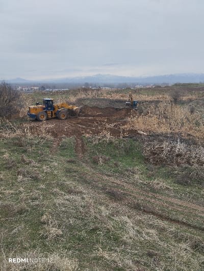 A yellow front loader is working on a dirt patch in a field, with a blue excavator visible further back. The landscape is a mix of dry grass and earth. Distant buildings and mountains are seen under a cloudy sky. A visible tire track leads through the foreground. The image is taken from an elevated perspective, showcasing the ongoing construction or land clearing activity.