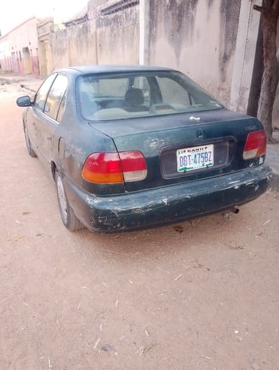 A dark green Honda Civic sits on a sandy, unpaved road. The car has a Kano license plate reading DBT-475BZ. Visible damage to the rear bumper suggests wear and tear. Behind the car is a tall, weathered concrete wall with a barbed wire fence on top. A tree trunk appears on the right side of the frame, close to the wall. In the distance, buildings line the opposite side of the street.