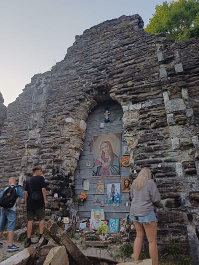 In Golovinka, Russia, an outdoor shrine is set within a large, weathered stone structure that appears to be a remnant of a larger building. The shrine is decorated with numerous religious icons, images, and crosses affixed to a wooden plank backing. The icons depict figures such as the Virgin Mary, Jesus Christ, and various saints. Smaller items, possibly votive offerings, are placed on shelves and ledges at the base of the shrine. Three individuals are present. Two young males stand to the left, facing the shrine. One wears a backpack and shorts, while the other has a black shirt and shorts. A blonde-haired woman is positioned to the right of the shrine, facing away from the camera and towards the religious display. The environment is rocky with some vegetation growing around the stone structure.